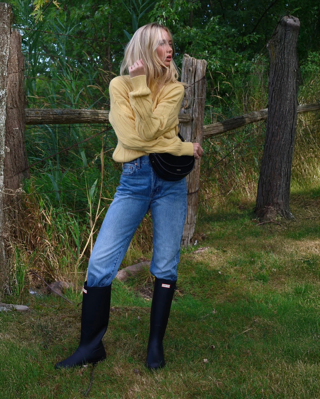 Woman in a yellow cable knit sweater standing next to a tree with jeans and hunter boots