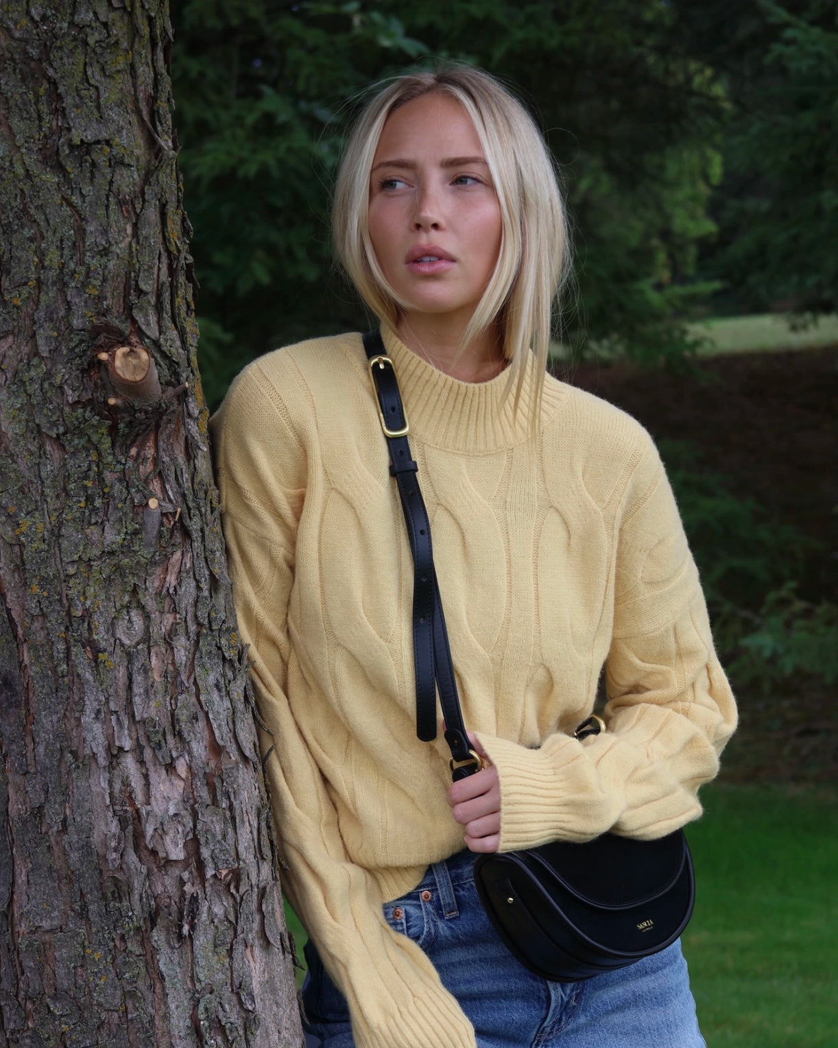 Woman in a yellow cable knit sweater standing next to a tree