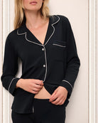 A woman wearing a black long pajama set with a button-up top and pants with light pink piping