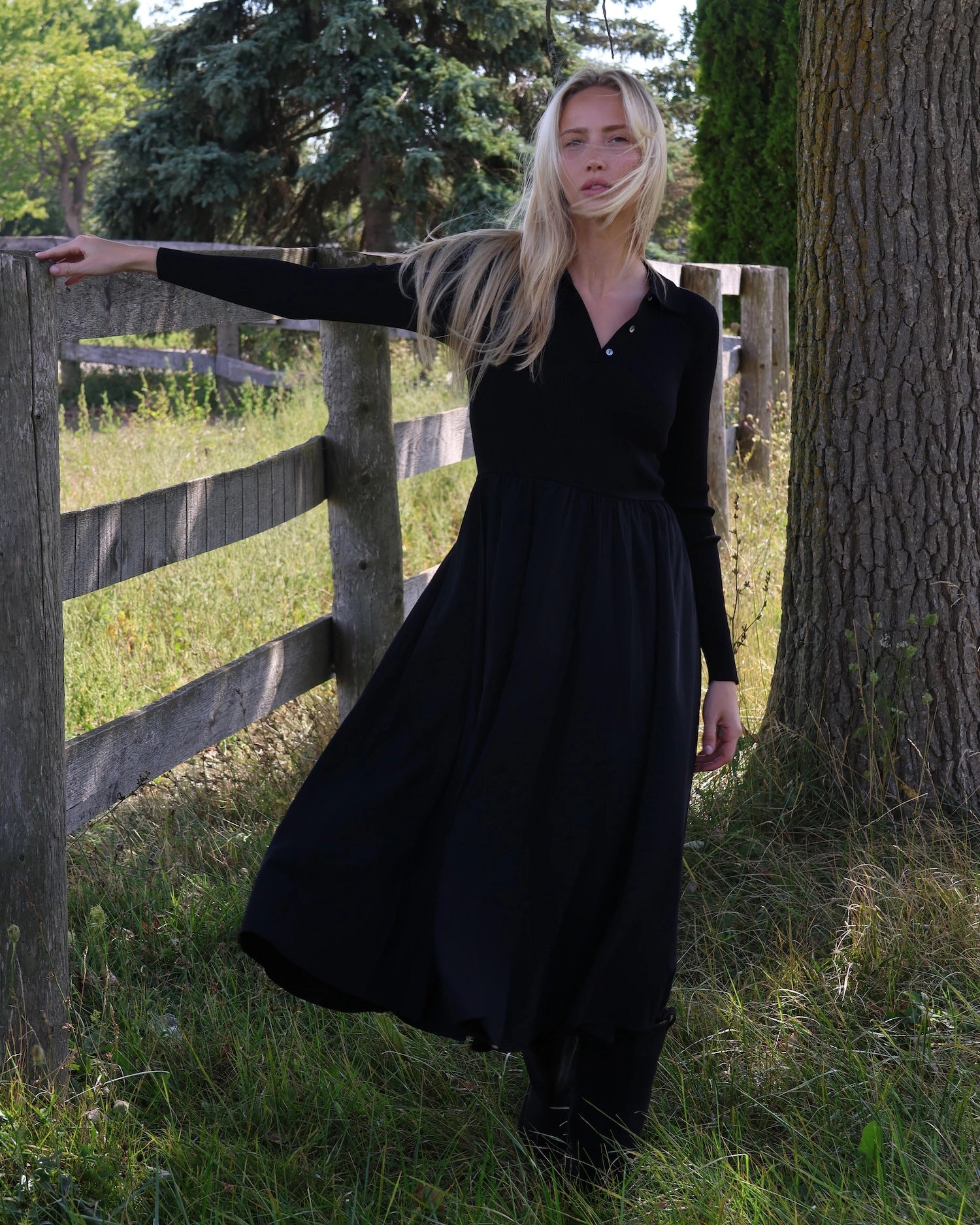 Woman in a long sleeve black dress with buttons and collar standing next to a wooden fence in a natural setting