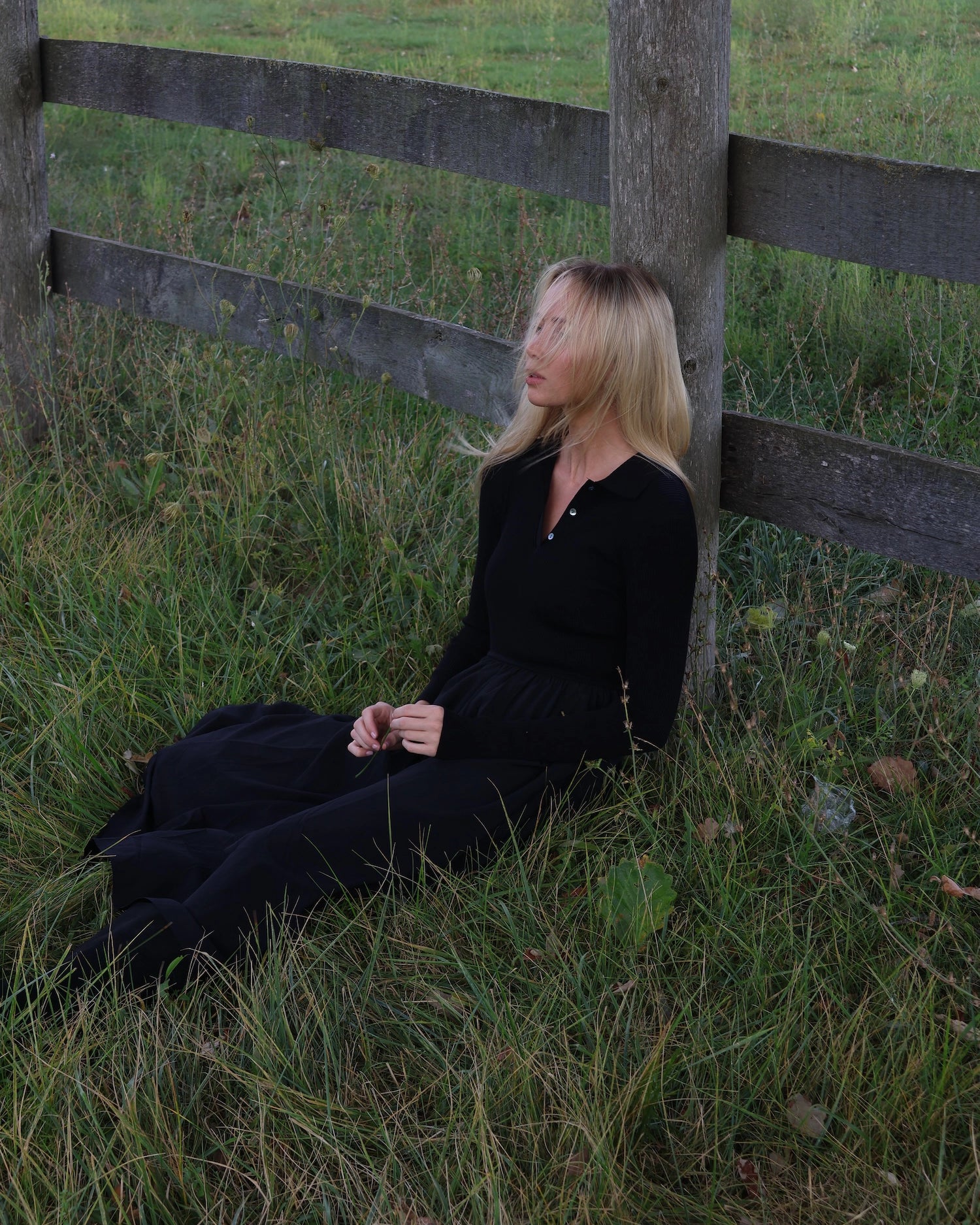Woman in a long sleeve black dress with buttons and collar standing next to a wooden fence in a natural setting