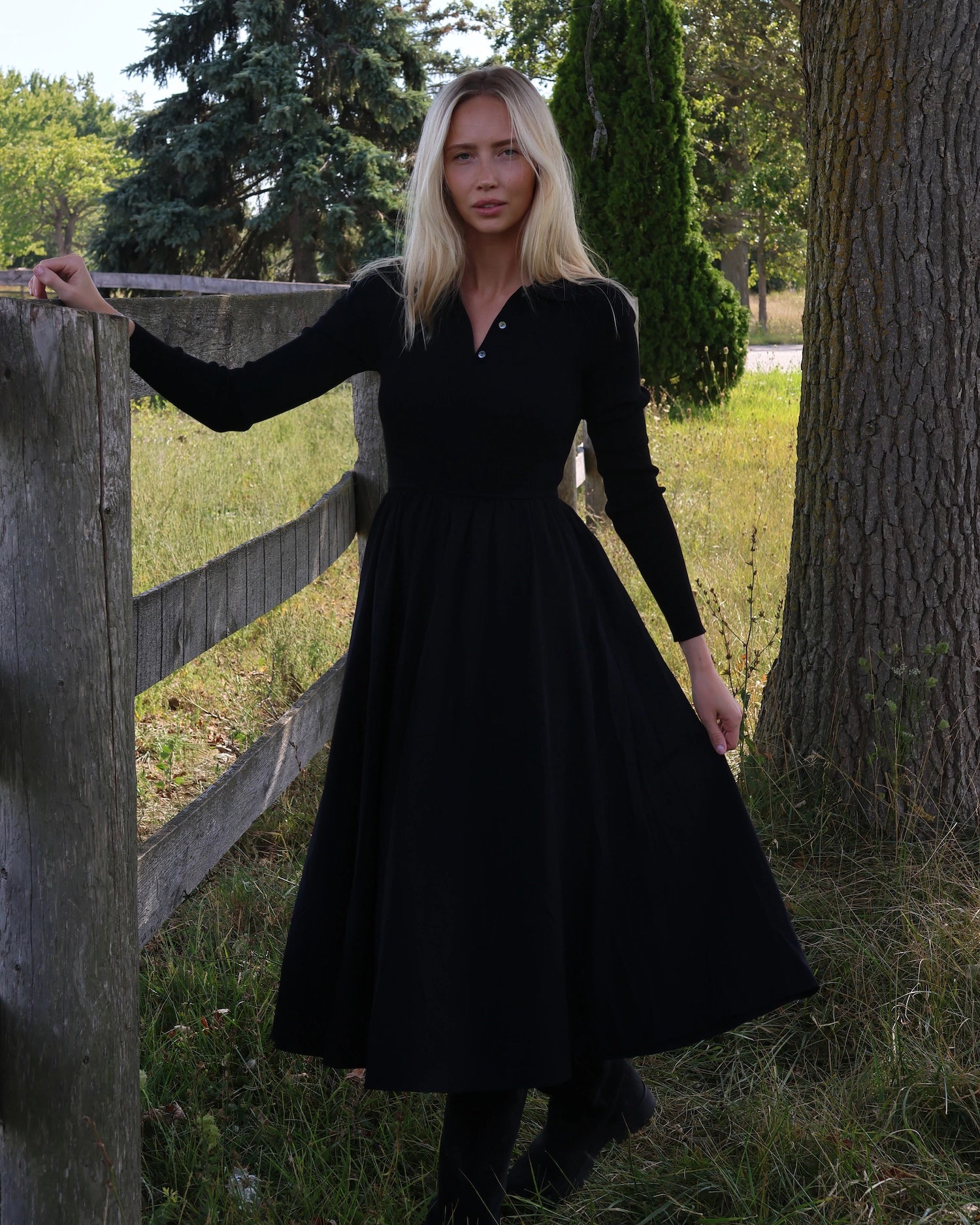 Woman in a long sleeve black dress with buttons and collar standing next to a wooden fence in a natural setting