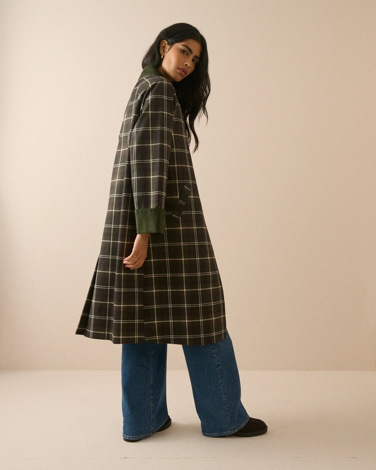 A model stands against a plain beige studio background wearing a long-line plaid coat with a dark brown base, cream-colored grid lines, and solid dark green collar, cuffs, and button accents