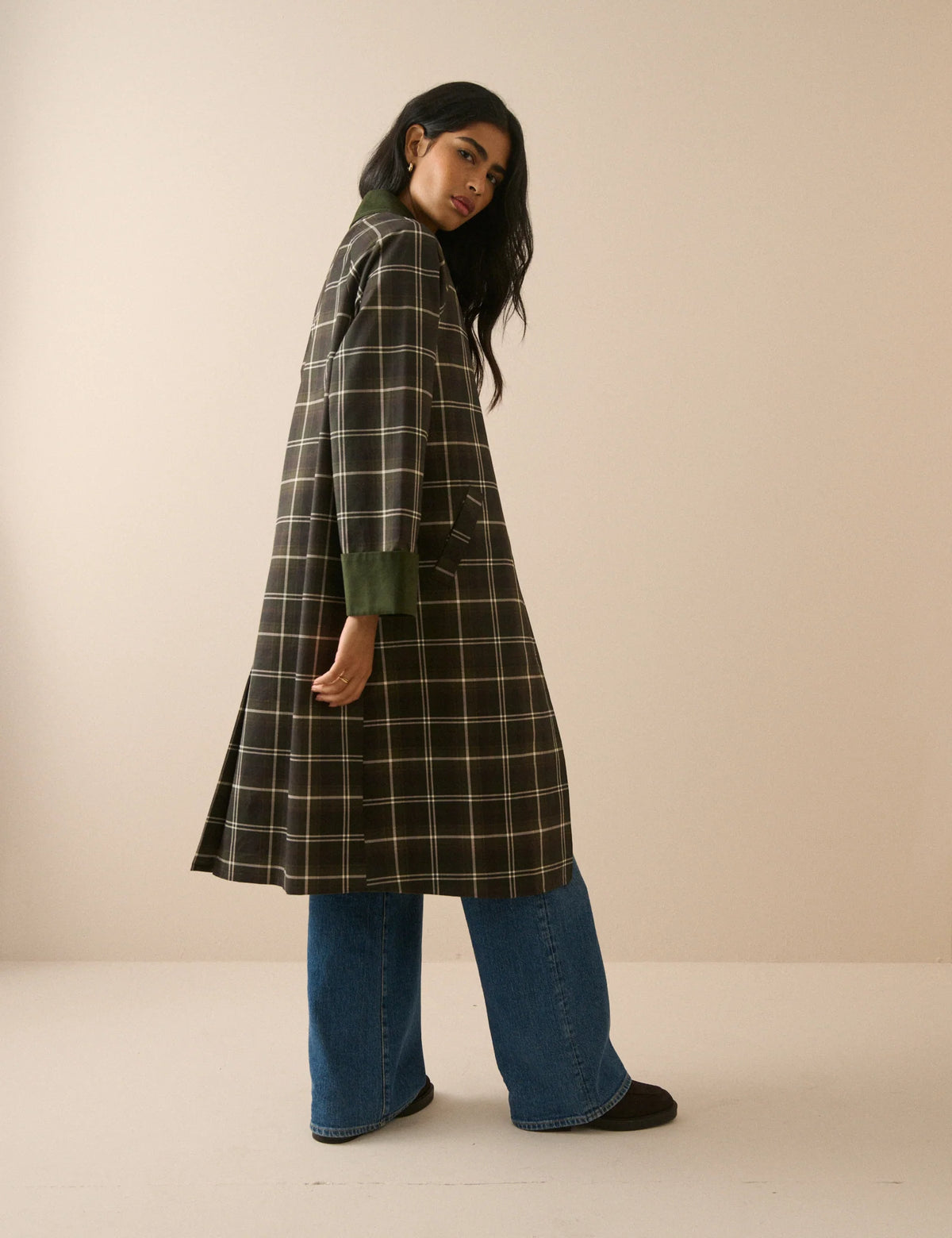 A model stands against a plain beige studio background wearing a long-line plaid coat with a dark brown base, cream-colored grid lines, and solid dark green collar, cuffs, and button accents