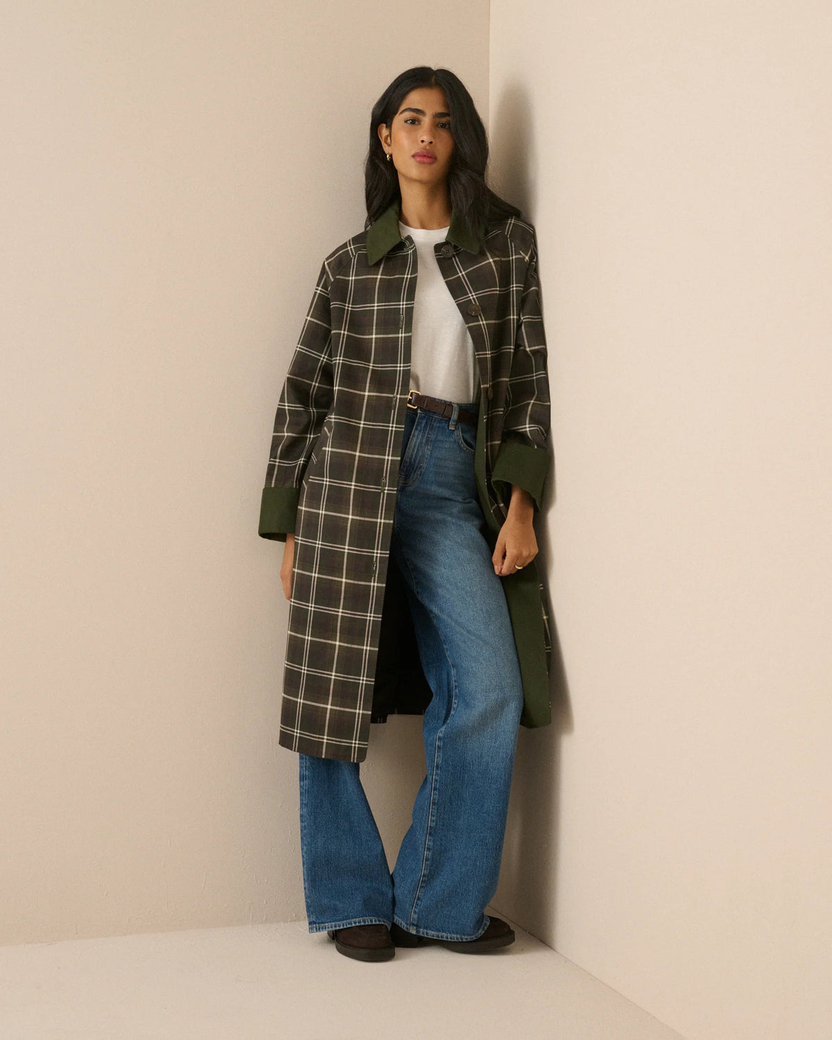A model stands against a plain beige studio background wearing a long-line plaid coat with a dark brown base, cream-colored grid lines, and solid dark green collar, cuffs, and button accents