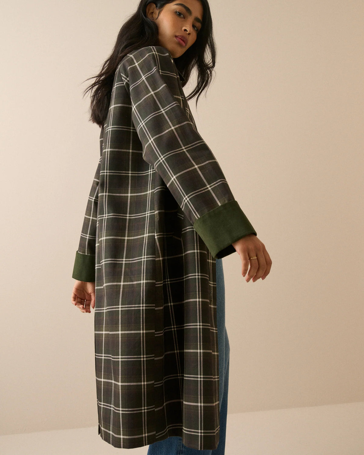 A model stands against a plain beige studio background wearing a long-line plaid coat with a dark brown base, cream-colored grid lines, and solid dark green collar, cuffs, and button accents