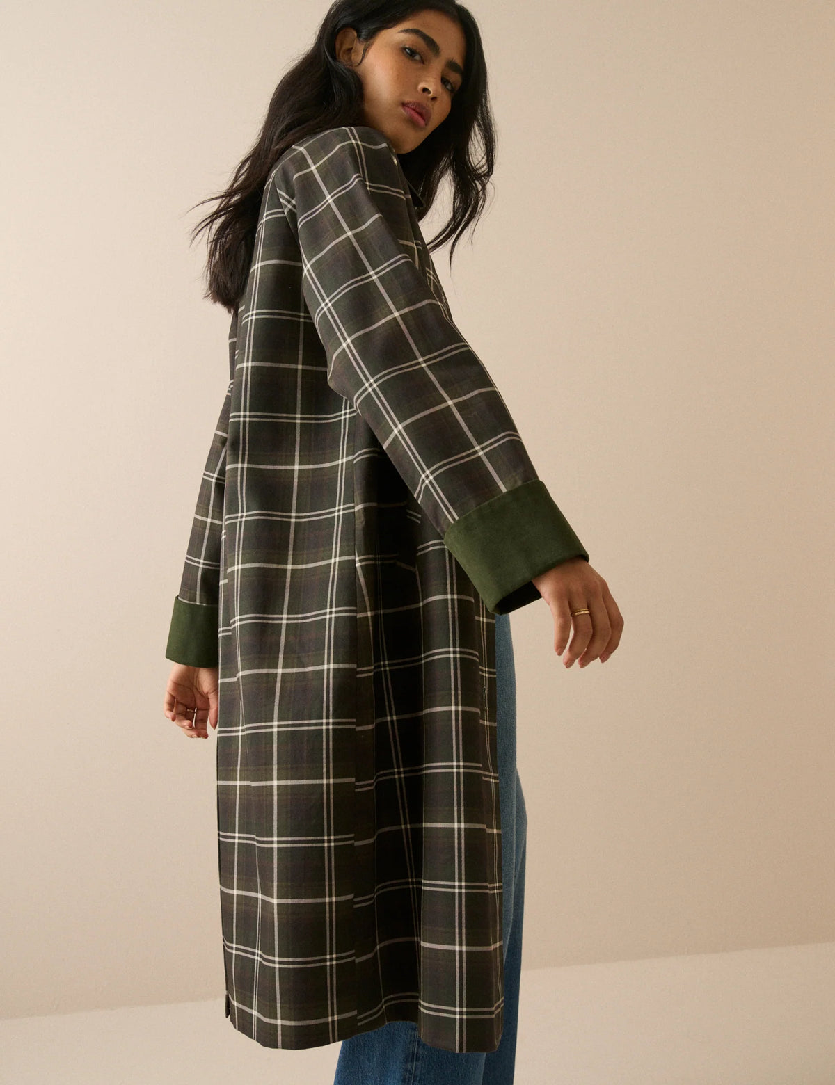 A model stands against a plain beige studio background wearing a long-line plaid coat with a dark brown base, cream-colored grid lines, and solid dark green collar, cuffs, and button accents