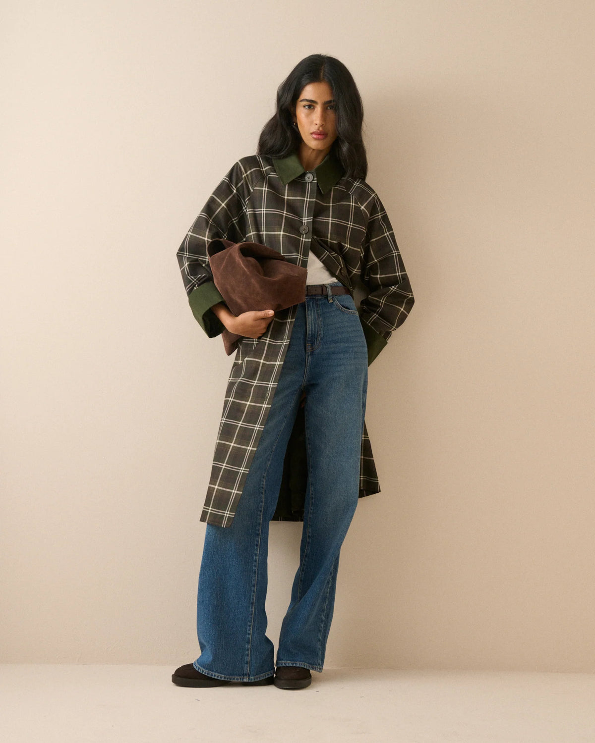 A model stands against a plain beige studio background wearing a long-line plaid coat with a dark brown base, cream-colored grid lines, and solid dark green collar, cuffs, and button accents