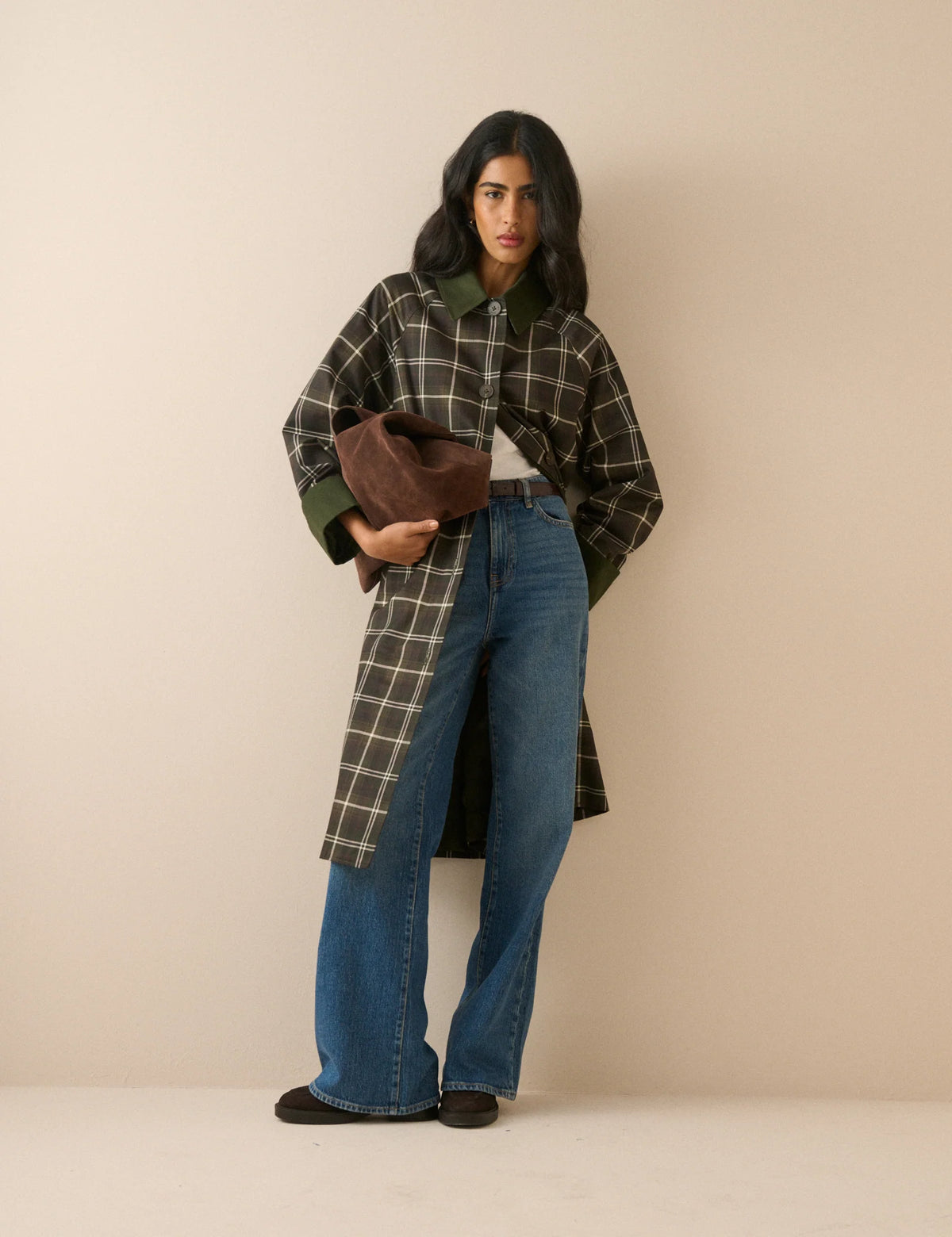 A model stands against a plain beige studio background wearing a long-line plaid coat with a dark brown base, cream-colored grid lines, and solid dark green collar, cuffs, and button accents
