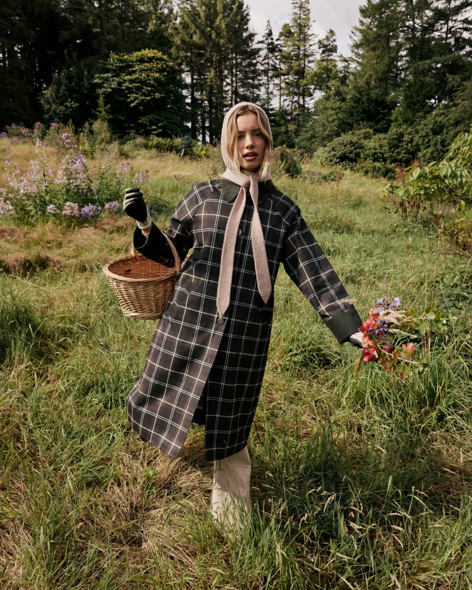 A woman stands in a grassy field holding a wicker basket, wearing a long green and white plaid coat with a wide beige collar and matching scarf, black gloves, and a beige head covering.