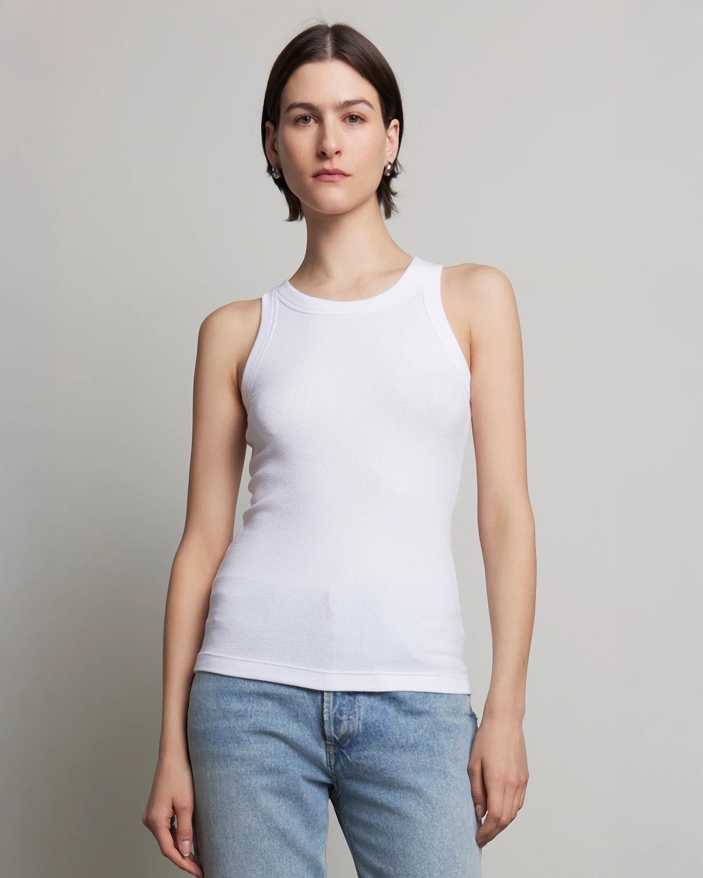 A woman wearing a white ribbed tank top and blue jeans.