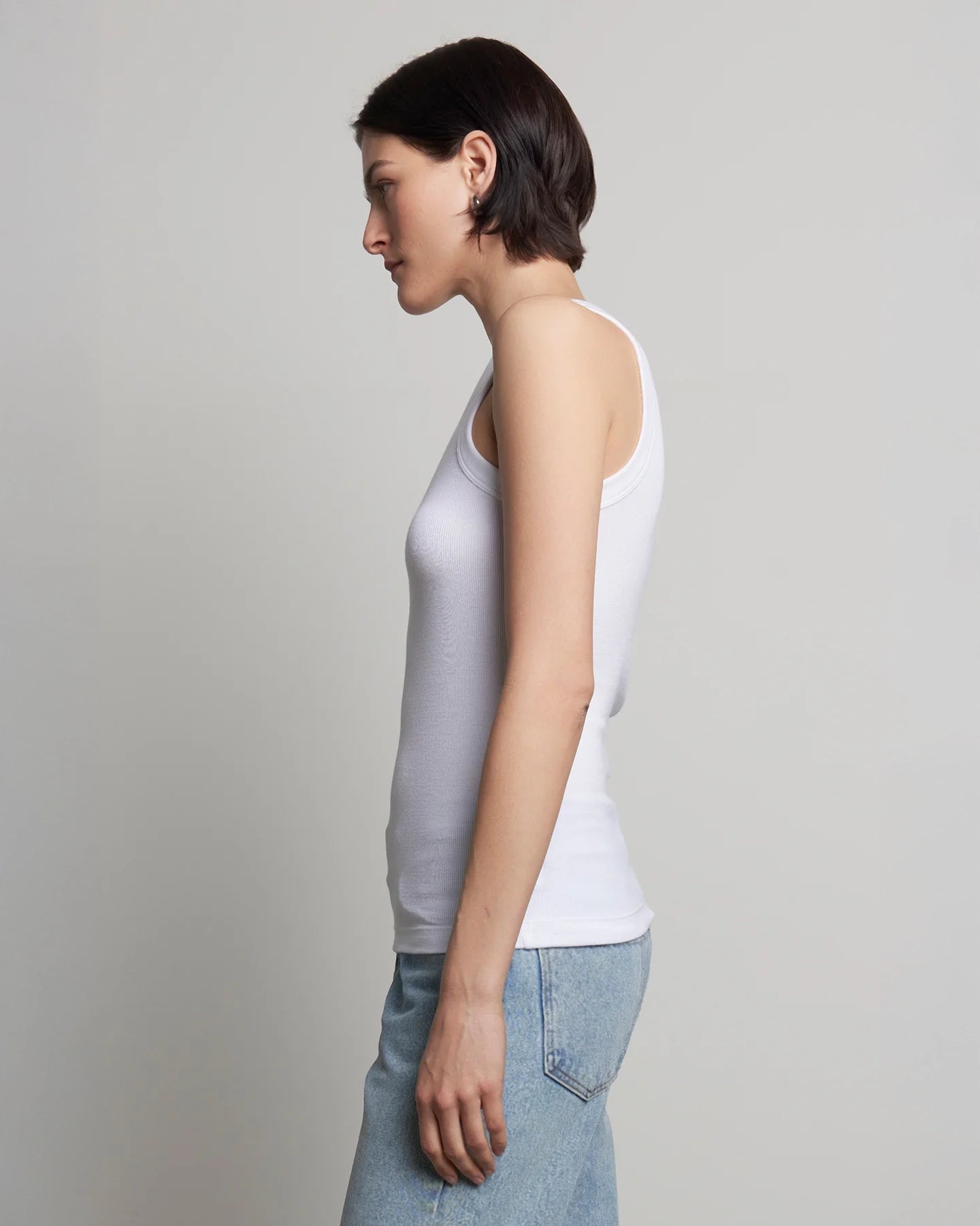 A woman wearing a white ribbed tank top and blue jeans.