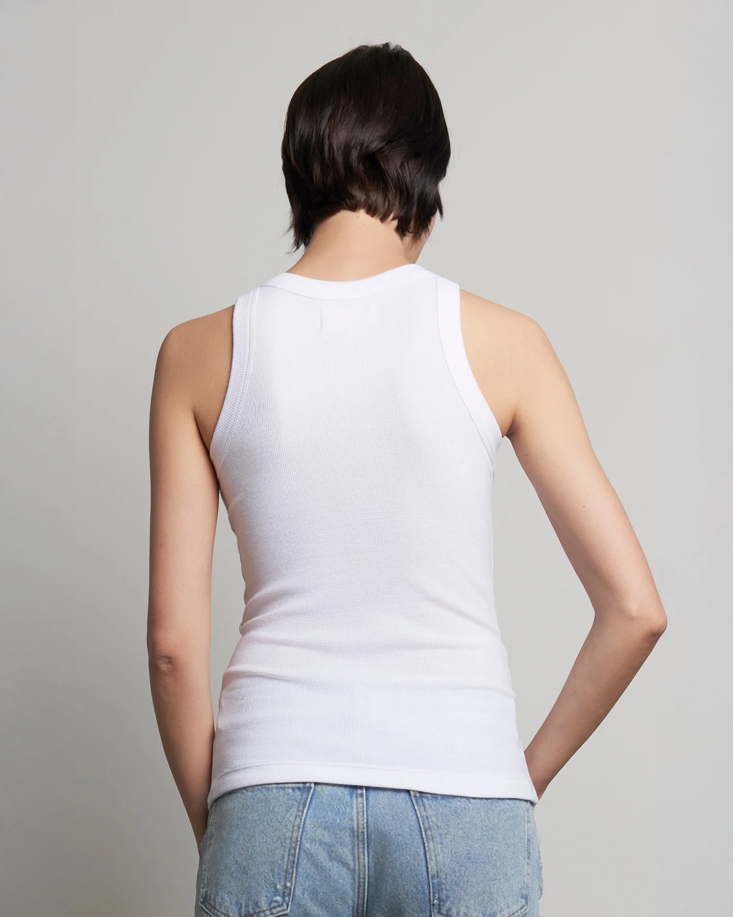 A woman wearing a white ribbed tank top and blue jeans.