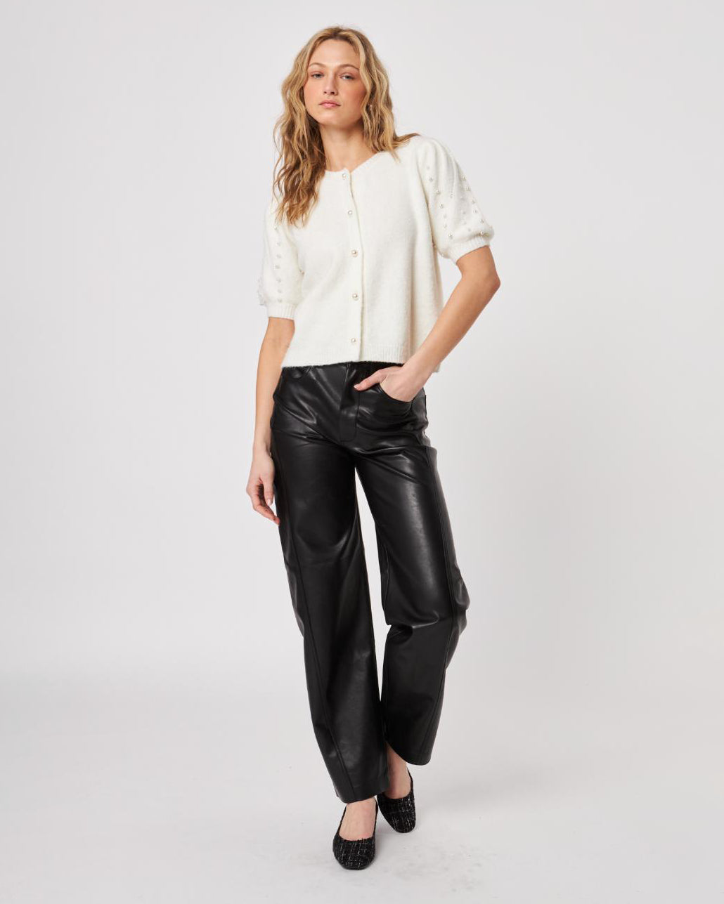 Woman wearing a white fluffy button down short sleeve cardigan with puff sleeves that have pearl embellishment on them and leather pants