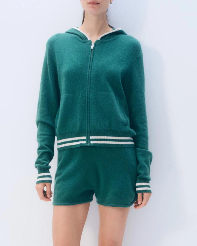 A woman wears a green varsity tracksuit style zip up hoodie with white stripes and matching shorts