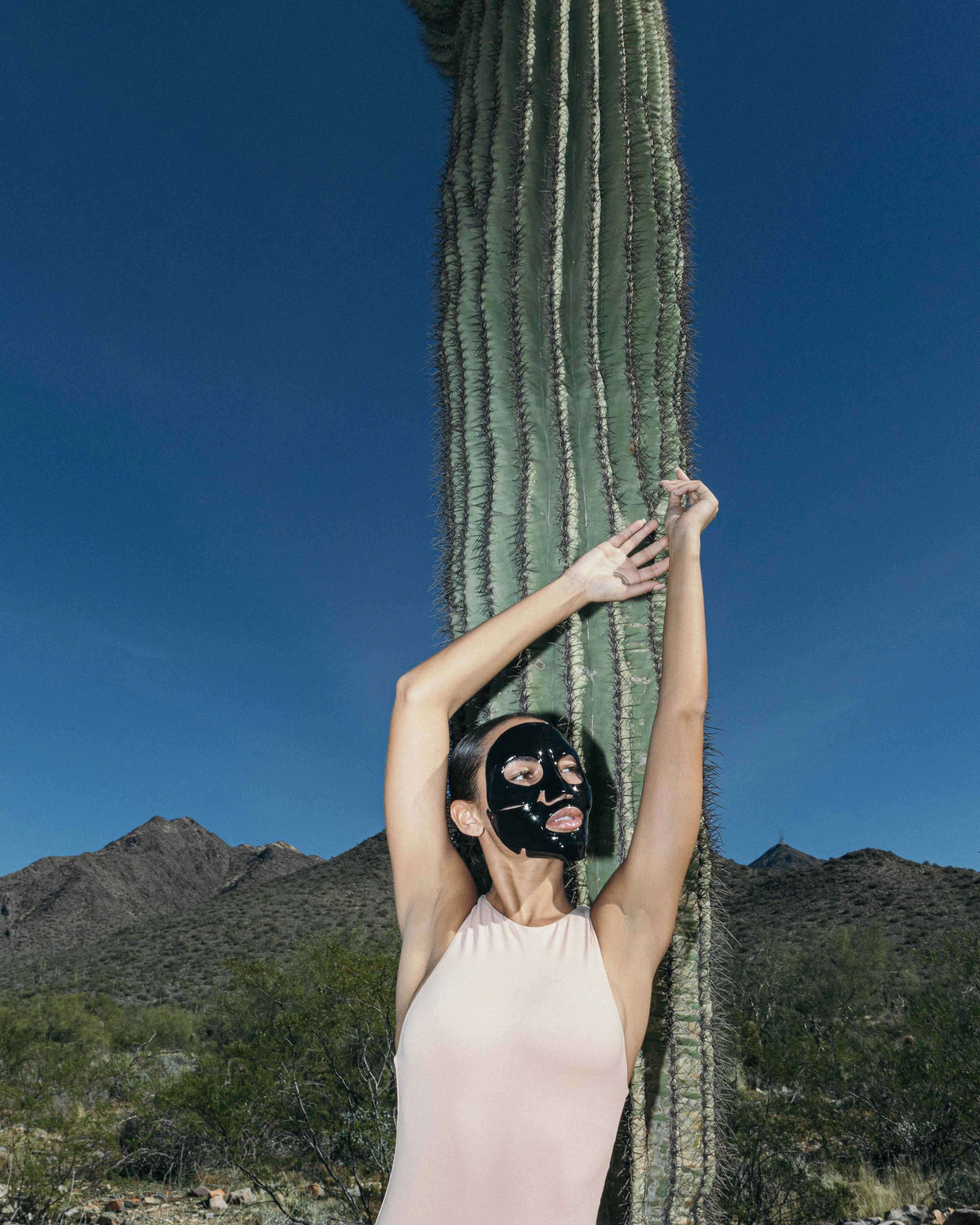 Person with a black mask standing next to a cactus in a desert landscape