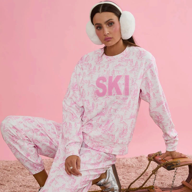 Woman wearing a pink and white 'SKI' sweatshirt and pants with white ear muffs by the brand Beach Riot