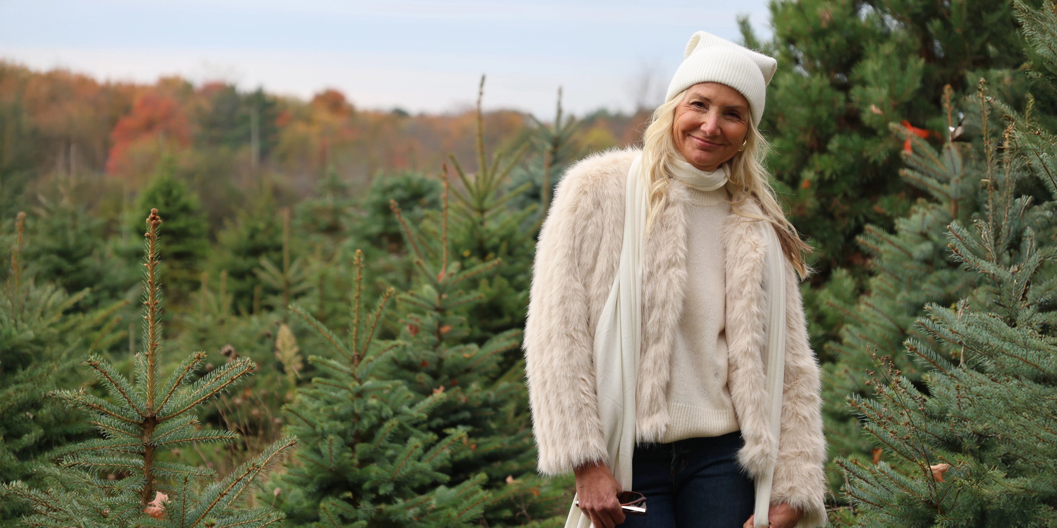 Woman in a white faux fur sweater and beanie hat standing amongst Christmas trees in a forest.