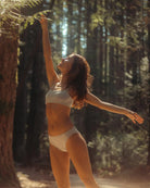 Woman in a grey modal bikini underwear and matching bra standing in a forest with sunlight filtering through the trees.