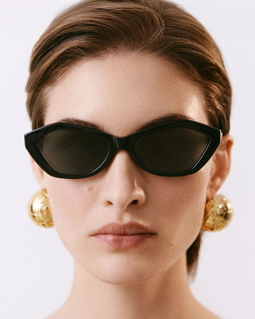 Woman wearing Jaquemus Bambino Angular Sunglasses in Black with Gold Details and big gold earrings