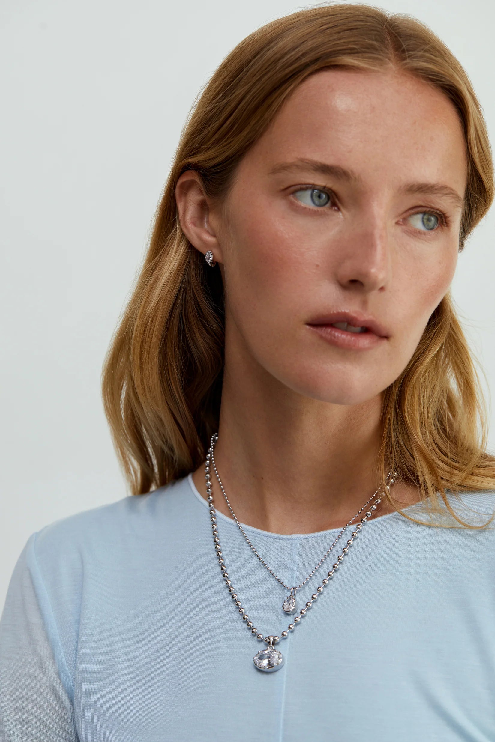 A woman wearing layered necklaces featuring a silver rhodium chain necklace with a big oval crystal pendant by brand Lili Claspe