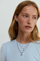 A woman wearing layered necklaces featuring a silver rhodium chain necklace with a big oval crystal pendant by brand Lili Claspe
