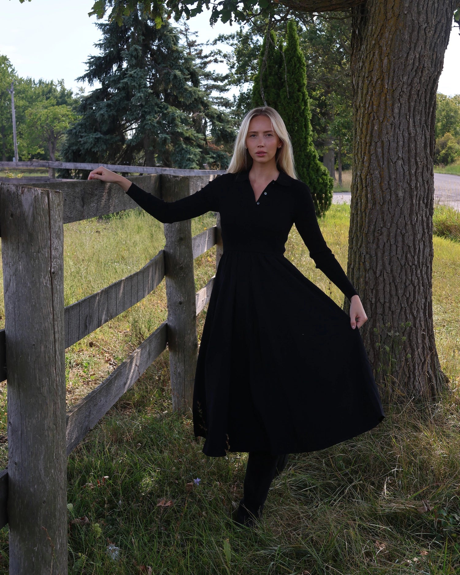 Woman in a long sleeve black dress with buttons and collar standing next to a wooden fence in a natural setting