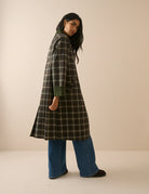 A model stands against a plain beige studio background wearing a long-line plaid coat with a dark brown base, cream-colored grid lines, and solid dark green collar, cuffs, and button accents