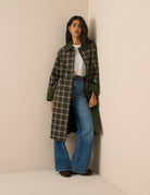 A model stands against a plain beige studio background wearing a long-line plaid coat with a dark brown base, cream-colored grid lines, and solid dark green collar, cuffs, and button accents