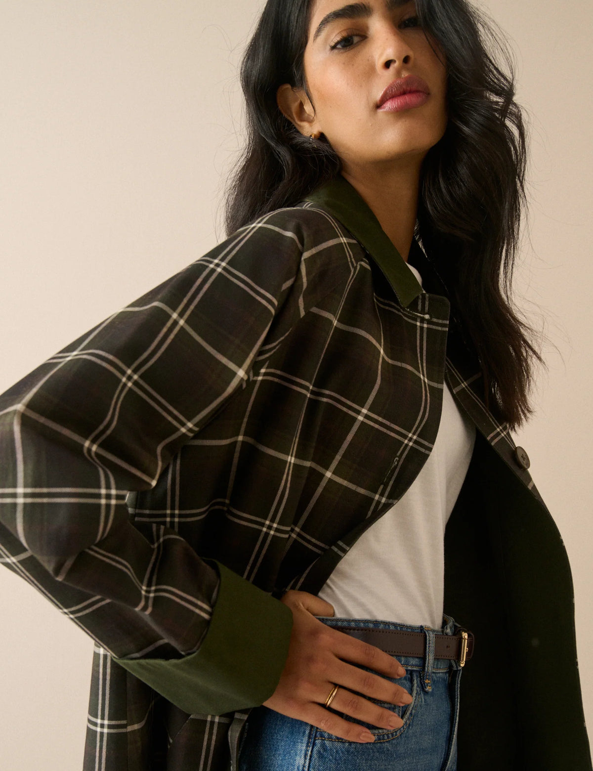 A model stands against a plain beige studio background wearing a long-line plaid coat with a dark brown base, cream-colored grid lines, and solid dark green collar, cuffs, and button accents