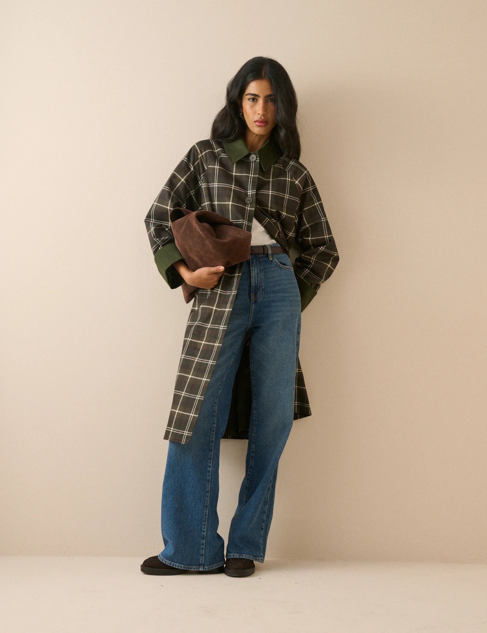 A model stands against a plain beige studio background wearing a long-line plaid coat with a dark brown base, cream-colored grid lines, and solid dark green collar, cuffs, and button accents