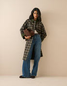 A model stands against a plain beige studio background wearing a long-line plaid coat with a dark brown base, cream-colored grid lines, and solid dark green collar, cuffs, and button accents
