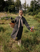 A woman stands in a grassy field holding a wicker basket, wearing a long green and white plaid coat with a wide beige collar and matching scarf, black gloves, and a beige head covering.
