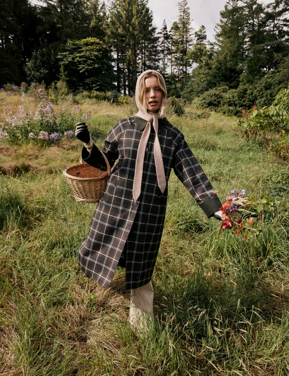 A woman stands in a grassy field holding a wicker basket, wearing a long green and white plaid coat with a wide beige collar and matching scarf, black gloves, and a beige head covering.