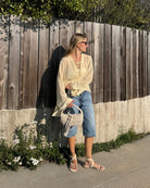Woman in a light blouse and denim shorts standing against a wooden fence with Beige ecru suede sandal with thin straps and gold toe detail