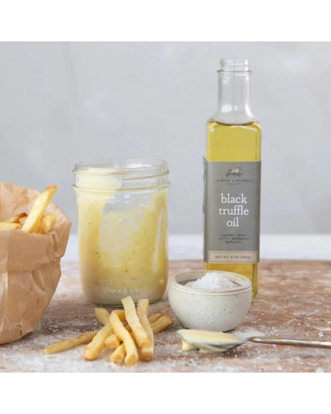 Bottle of black truffle oil with a jar of dressing, fries, and salt on a wooden surface.