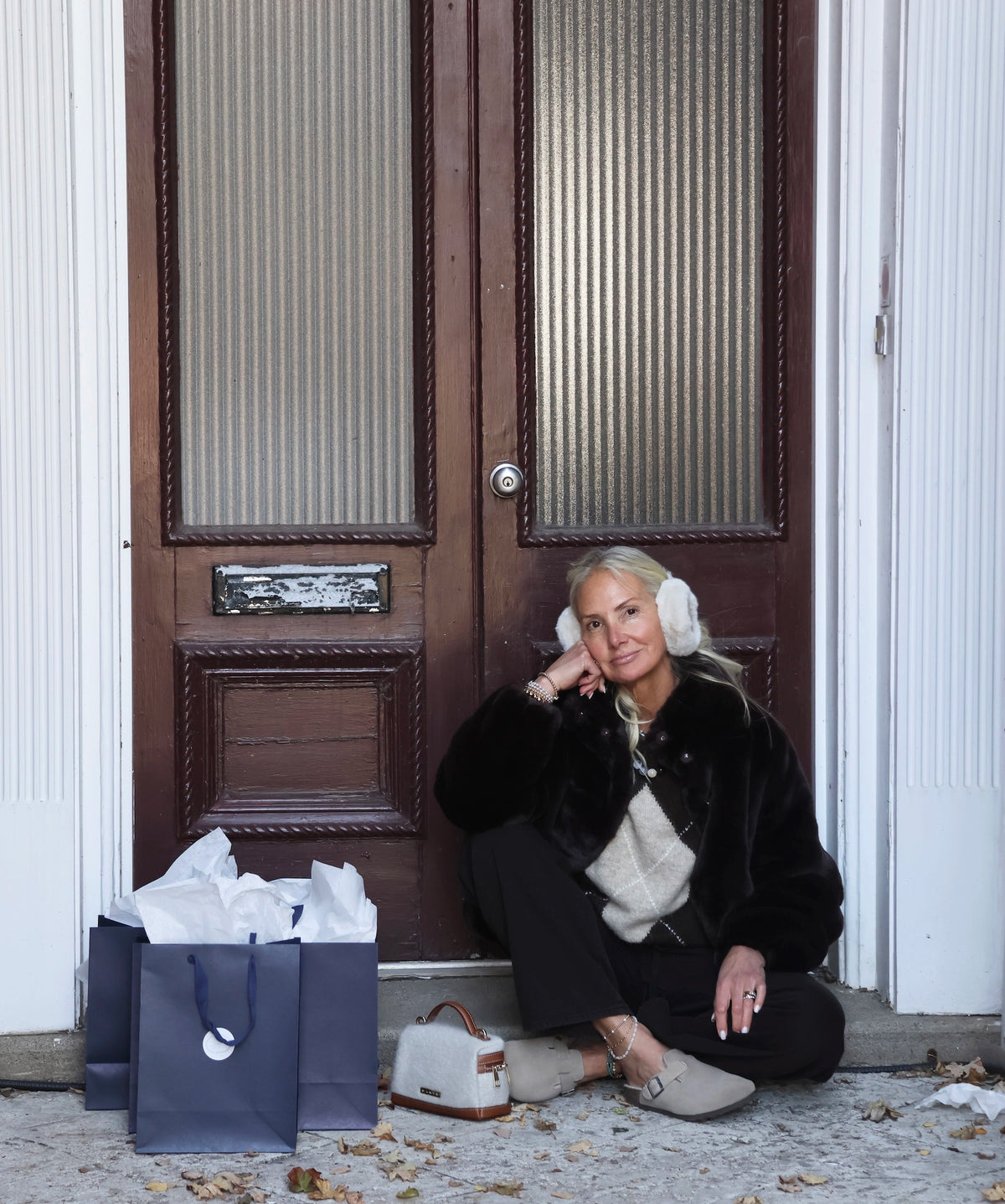 A woman sits on the steps of a brown wooden door wearing a dark brown faux-fur jacket, cream argyle knit sweater, black pants, and beige slip-on shoes, with white earmuffs on. A navy shopping bag and a small beige handbag sit beside her on the ground.