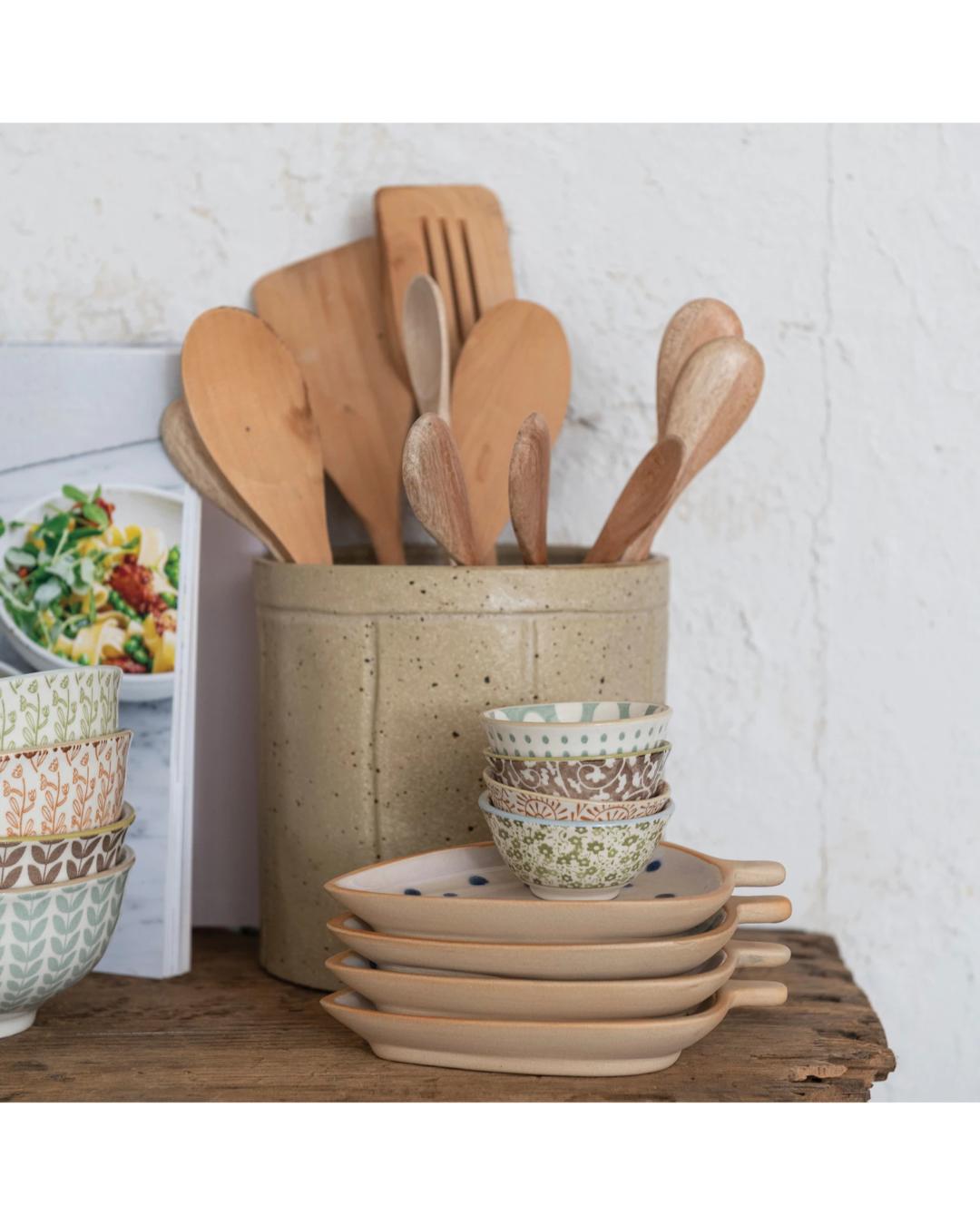 Set of ceramic bowls and wooden utensils on a wooden surface with a white tiled wall background.