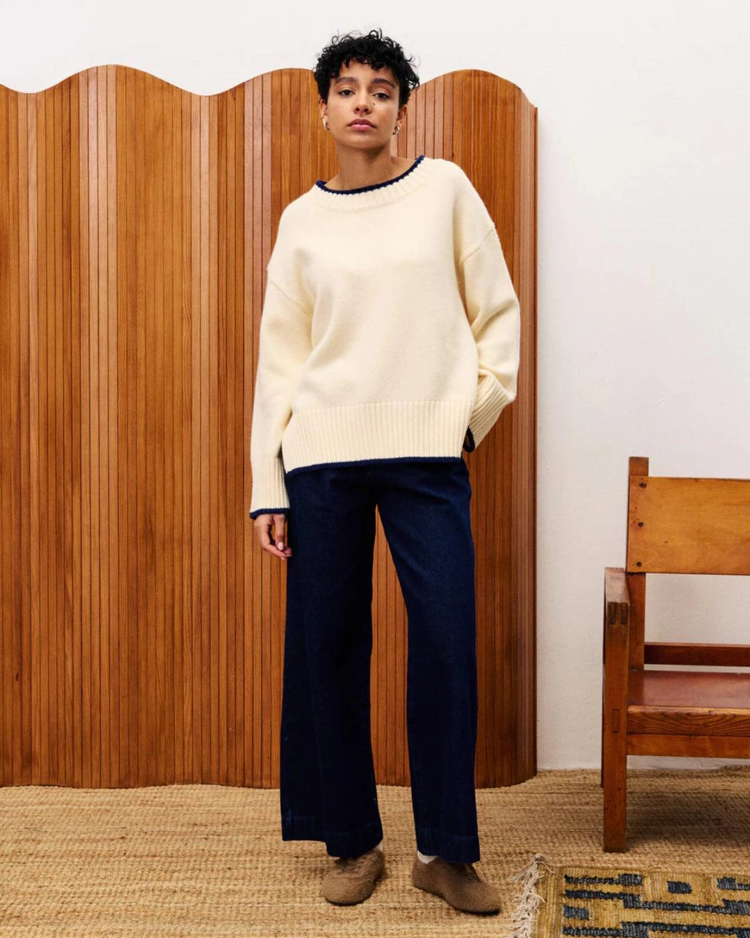 Person wearing a cream sweater and dark pants standing in front of a wooden screen.